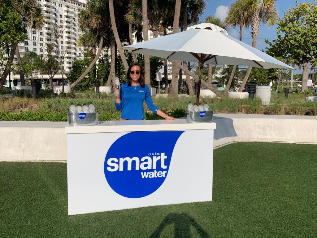A female brand ambassador holding a Smartwater bottle and standing behind an event table.